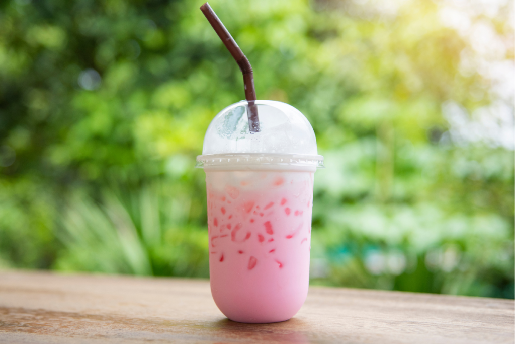 A refreshing pink drink served in a clear plastic cup with ice, topped with a dome lid and a brown straw, placed on a wooden table with a green blurred background.