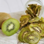 A glass jar spilling dehydrated kiwi slices onto a white wooden board next to a whole and a sliced fresh kiwi.